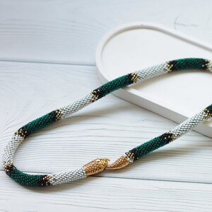 Pode incluir: Um colar de contas brancas, verdes e douradas com um fecho de cobra. O colar está sobre uma superfície branca.