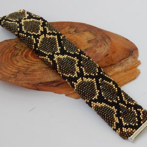 May include: A black and gold beaded bracelet with a snake skin pattern. The bracelet is on a wooden surface.
