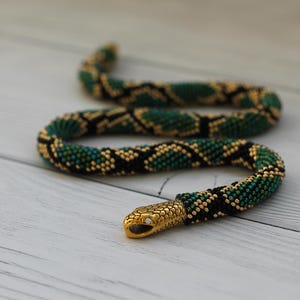 May include: A flexible snake-shaped accessory crafted from tiny beads in shades of emerald green, black, and gold. The body features a repeating diamond pattern, while the head is made of polished gold-tone metal with a small clear stone for an eye.