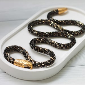 Pode incluir: Um colar de cobra de contas pretas e douradas com um fecho de cabeça de cobra dourada. O colar é apresentado num prato oval branco.
