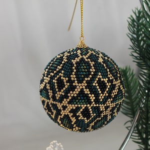 Może przedstawiać: Ozdoba choinkowa z koralików z geometrycznym wzorem. Ozdoba ma kształt kuli, głównie ciemnozielonej z złotym wzorem. Wisi na złotym łańcuszku i znajduje się obok zielonej gałęzi sosny.