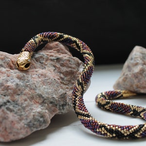 Puede incluir: Un collar de serpiente de cuentas con una cabeza dorada y un esquema de color negro, dorado y morado. El collar está enrollado en una roca.