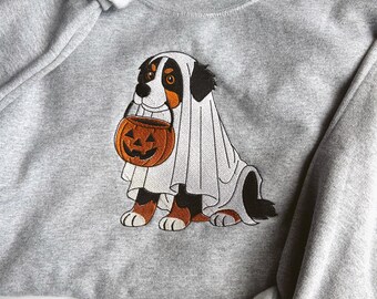 Puede incluir: Una sudadera gris con un diseño de Halloween. El gráfico muestra un perro disfrazado de fantasma, sosteniendo una calabaza naranja tallada. El perro tiene marcas negras, marrones y blancas. La sudadera tiene cuello redondo y mangas largas.