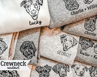 May include: A stack of gray and white crewneck sweatshirts with custom dog portraits and names printed on them. The names include "Lucky", "Charlie + Willow", "Charlie", "Georgie", "Baxter + Kai", "Ellie + Belle".