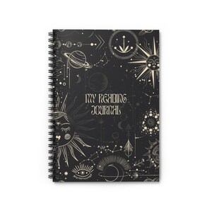 Könnte beinhalten: Ein schwarzes Spiralnotizbuch mit einem himmlischen Design. Der Einband zeigt ein sich wiederholendes Muster aus Sonnen, Monden, Sternen und Planeten in Cremefarbe. Der Text "MY READING JOURNAL" ist in Creme auf dem Einband gedruckt.