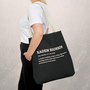 Könnte beinhalten: Schwarze Tragetasche mit cremefarbenen Trägern. Auf der Tasche steht in Weiß der Name "XADEN RIORSON" mit einer Definition. Die Definition beschreibt einen schattenwirkenden, drachenreitenden Revolutionär. Die Tasche wird über der Schulter getragen.