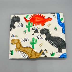 Puede incluir: Una tela blanca con un patrón repetido de dinosaurios y cactus. Los dinosaurios son de varios colores, incluyendo rojo, naranja, marrón, negro y azul. El texto "ROAR!" aparece dos veces en la tela.