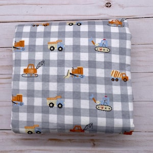 Puede incluir: Una manta plegada con un patrón de cuadros grises y blancos. La manta está decorada con vehículos de construcción naranjas, azules y blancos, incluyendo volquetes, excavadoras y excavadoras. La manta está hecha de un material suave.