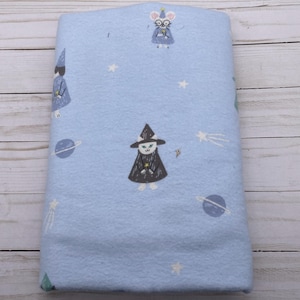 May include: A light blue fleece blanket featuring whimsical illustrations of cartoon wizards, mice, planets, stars, and shooting stars. The blanket is folded and ready for use. The design is repeated across the fabric.