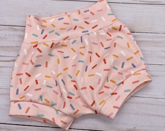 May include: Pink baby shorts with a colorful sprinkle pattern. The shorts have a high waistband and rolled cuffs. The sprinkles are in shades of red, blue, yellow, and white. The shorts are made of a soft fabric.