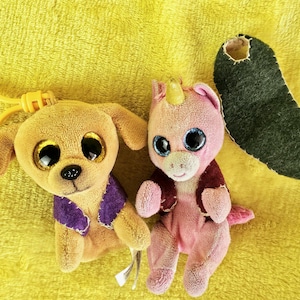 May include: A plush dog with a yellow keychain, a pink unicorn, and a green felt shape on a yellow background. The dog has large, gold-colored eyes and a purple vest. The unicorn has blue eyes and a red vest.