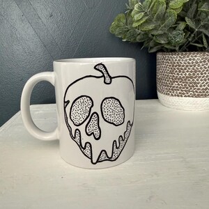 May include: White ceramic mug featuring a black line drawing of a skull-shaped apple. The design includes dotted details and a dripping effect. The mug has a curved handle and is set against a neutral background.