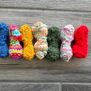 May include: Seven small, hand-knitted items arranged in a horizontal row. The colors include solid blue, a multi-colored yarn, solid yellow, a variegated tan and white, solid dark green, a variegated pink and white, and a solid bright red.