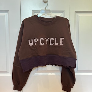 May include: A brown cropped sweatshirt featuring the word UPCYCLE in light pink block letters across the chest. The garment has long sleeves with ribbed cuffs and a darker brown horizontal band at the bottom with a raw, unfinished hemline.
