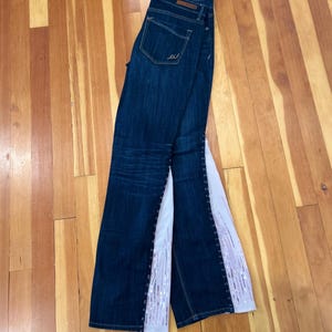 May include: Dark blue denim jeans featuring a high-waisted design with a back pocket and a vertical white fabric panel insert along the outer leg seams. The white panels are embellished with rows of shimmering silver sequins in a vertical pattern.