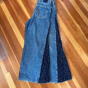 May include: A pair of blue denim jeans featuring a wide-leg silhouette with a contrasting dark navy blue fabric insert patterned with small white dots. The jeans include a standard waistband with belt loops, a back pocket, and a dark blue label.