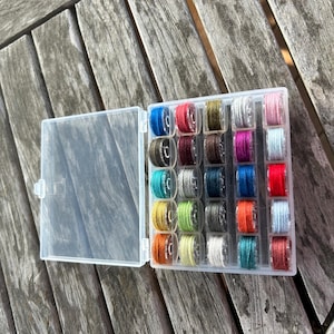 May include: A clear plastic box filled with spools of colorful thread. The box has a lid that is open, revealing the spools of thread. The spools are arranged in rows and columns, and they are all different colors.