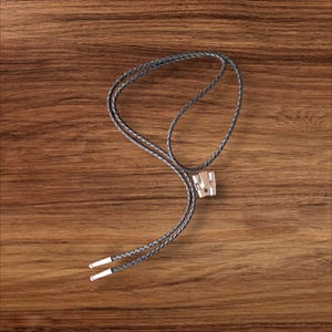 May include: A bolo tie with a silver-colored metal slide and braided black leather cord. The cord ends have silver-colored metal tips. The tie is displayed on a wooden surface.