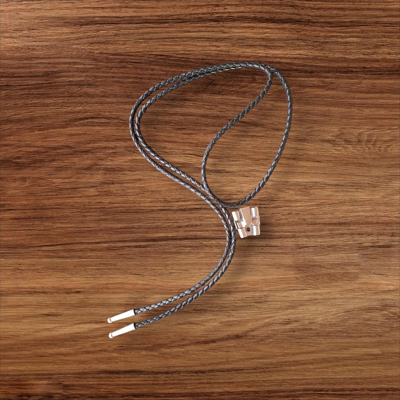 May include: A bolo tie with a silver-colored metal slide and braided black leather cord. The cord ends have silver-colored metal tips. The tie is displayed on a wooden surface.