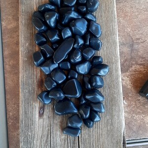 May include: A collection of smooth, polished black stones arranged on a weathered wooden plank. The stones vary in size and shape, reflecting light. The wood has a natural, aged appearance, with visible grain and knots.