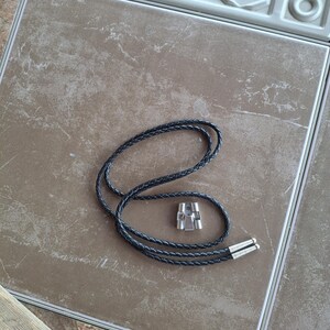 May include: A black braided leather bolo tie with silver-colored metal tips and a silver-colored metal slide. The tie is lying on a brown tile surface.