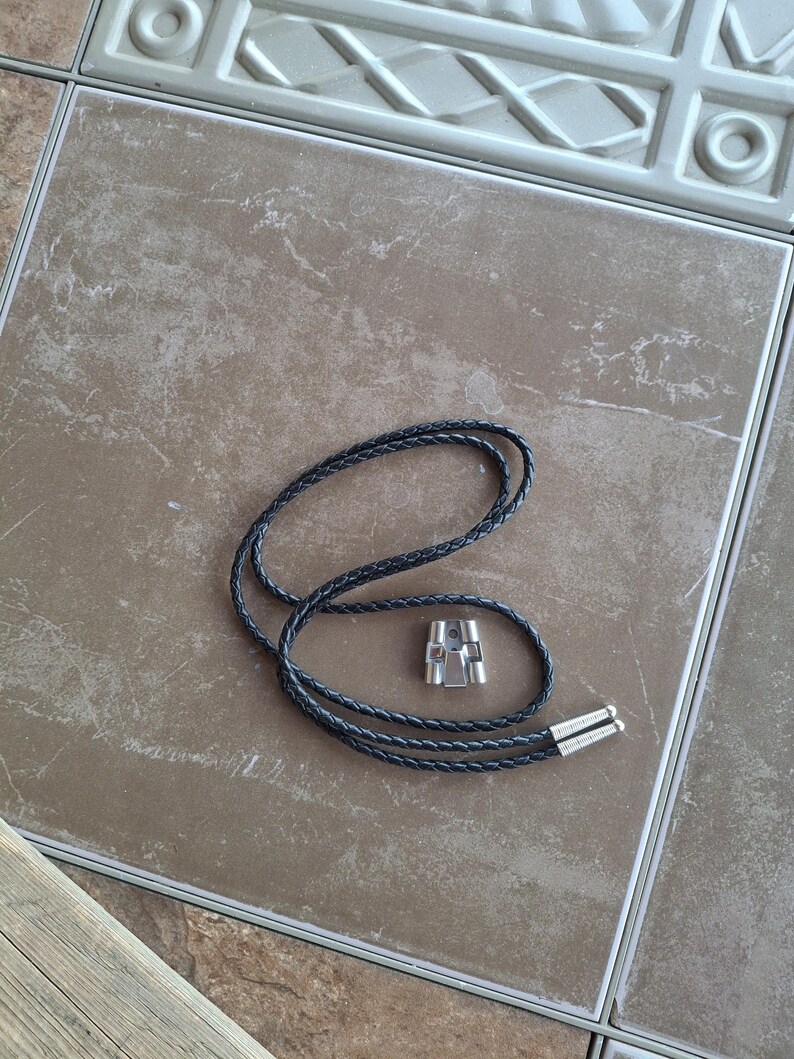 May include: A black braided leather bolo tie with silver-colored metal tips and a silver-colored metal slide. The tie is lying on a brown tile surface.