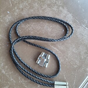 May include: A black braided leather bolo tie with a silver-colored metal slide. The tie has a braided design and silver-colored metal tips. The slide has a geometric design.