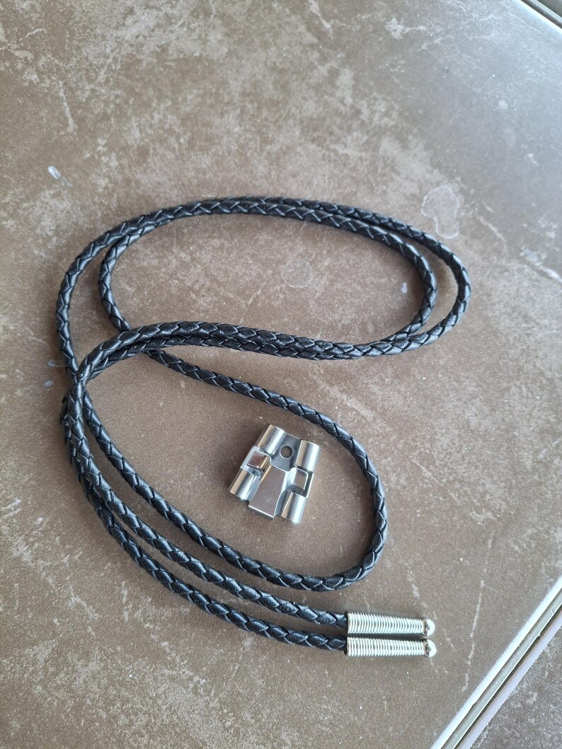 May include: A black braided leather bolo tie with a silver-colored metal slide. The tie has a braided design and silver-colored metal tips. The slide has a geometric design.