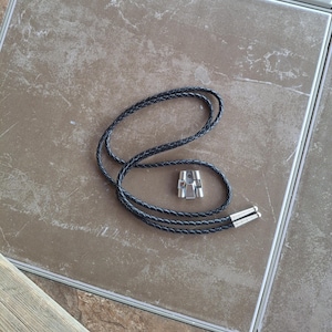 May include: A black braided leather bolo tie cord featuring a silver-toned metal slide clasp and two matching silver-toned metal aglets at the ends of the cord. The cord is arranged in a loose loop, showcasing the texture of the braided material.