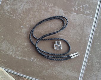 May include: A black braided cord bolo tie featuring a silver-toned metal slide clasp and matching silver-toned metal aglets at the ends of the cord. The cord is arranged in a loose loop, showcasing the braided texture and the metallic hardware components.