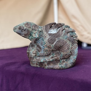 May include: A carved stone sculpture of a lizard in a curled position. The sculpture features a mottled brown and teal green color palette with visible textured scales and defined ridges along the back, showcasing the natural patterns of the stone.
