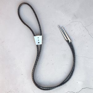 May include: A bolo tie featuring a black braided leather cord with a silver-toned metal slide clasp. The clasp has a trapezoidal shape with three small circular cutouts. The cord ends are finished with two pointed silver-toned metal tips.