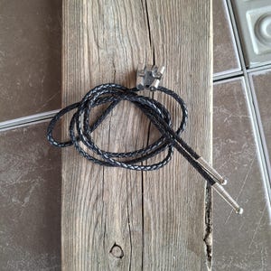 May include: A black leather bolo tie with silver-colored metal tips and a decorative clasp. The tie is coiled on a weathered, gray wooden plank, showcasing the texture of the wood.