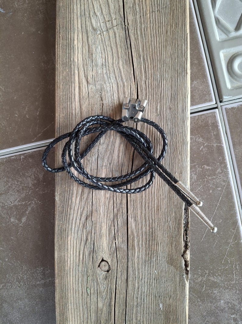 May include: A black leather bolo tie with silver-colored metal tips and a decorative clasp. The tie is coiled on a weathered, gray wooden plank, showcasing the texture of the wood.