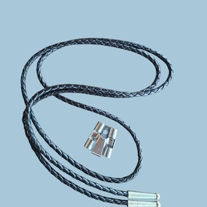 May include: A black braided leather bolo tie with silver-colored metal tips and a decorative clasp. The tie is displayed against a light blue background. The clasp has a rectangular shape with a geometric design.