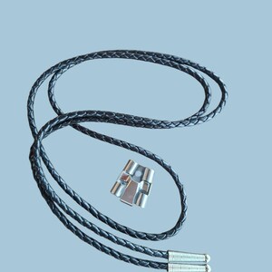 May include: A black braided leather bolo tie with a silver-colored metal clasp. The tie features a woven design and silver tips at the ends. The clasp has a rectangular shape with a geometric design.