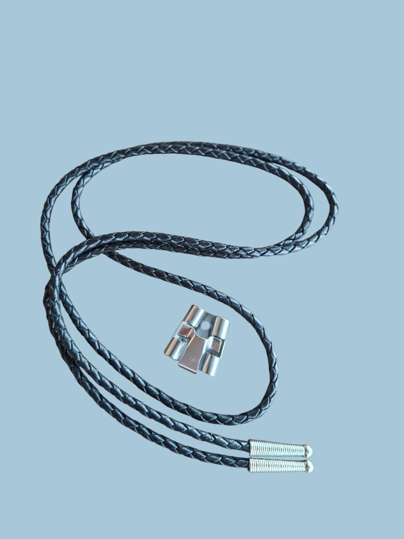 May include: A black braided leather bolo tie with a silver-colored metal clasp. The tie features a woven design and silver tips at the ends. The clasp has a rectangular shape with a geometric design.