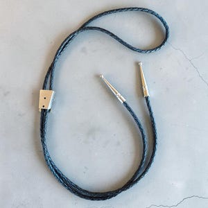 May include: A black braided bolo tie featuring a silver-toned metal slide clasp and two matching silver-toned metal aglets at the ends of the cord. The cord is constructed from a woven material and forms a continuous loop with a sliding adjustment.