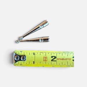 May include: Two silver-toned metal aglets with a tapered, conical shape and a small spherical tip. The aglets are positioned above a yellow Stanley measuring tape, which indicates that each piece is approximately 1.75 inches in length.