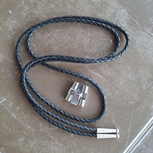 May include: A black braided leather bolo tie with a silver-colored metal clasp. The tie features a woven design and silver tips at the end of the cord. The clasp has a geometric design.