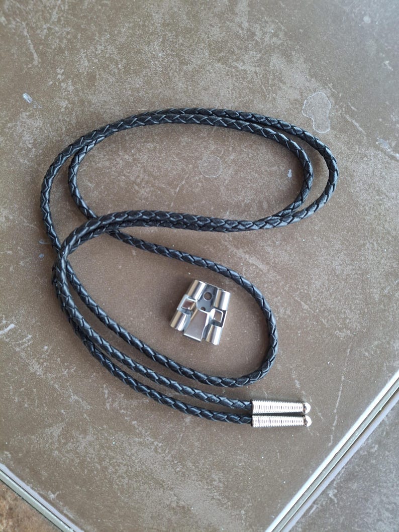 May include: A black braided leather bolo tie with a silver-colored metal clasp. The tie features a woven design and silver tips at the end of the cord. The clasp has a geometric design.