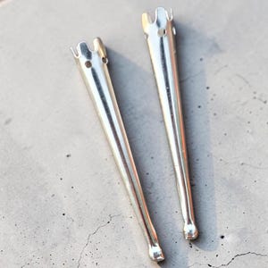 May include: Two silver-colored metal candle snuffers with a tapered, conical shape. Each snuffer features a crown-like top with small circular cutouts and a rounded, ball-shaped tip at the narrow end, showcasing a smooth, reflective metallic finish.