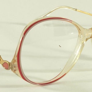 May include: A pair of clear plastic eyeglasses with a red trim. The frames have a unique, curved shape and the brand name "JORDACHE" is printed on the right temple.