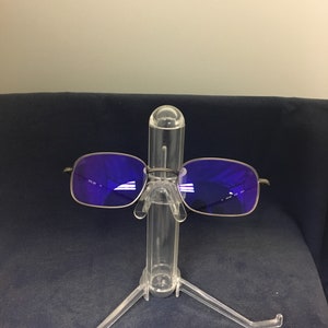 May include: A pair of square-shaped sunglasses with a silver frame and purple lenses. The sunglasses are displayed on a clear plastic stand.