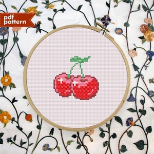 May include: A cross-stitch pattern of two red cherries with green stems and leaves on a pink background. The pattern is in a wooden embroidery hoop.