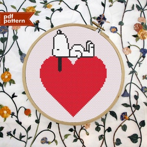 May include: A cross-stitch design in a wooden hoop shows a red heart with a white figure resting on it. The figure has a black nose and is flexing a bicep. The text "pdf pattern" is in the upper left corner.