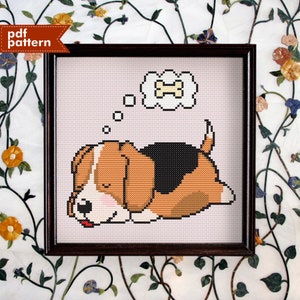 Könnte beinhalten: Kreuzstichmuster eines schlafenden Beagle-Hundes, der von einem Knochen träumt. Der Hund ist braun, schwarz und weiß. Der Knochen ist gelb. Das Muster ist in einem weißen Rahmen.