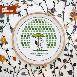 Op de afbeelding: Een kruissteekpatroon met Snoopy die een groene paraplu vasthoudt onder een regen van groene vierbladige klavers. Het patroon is ingelijst in een houten hoepel en bevat de tekst "Happy St. Patrick's Day".