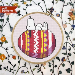 May include: A cross-stitch pattern of Snoopy, the iconic beagle from Peanuts, sleeping on a colourful, striped, red, yellow and white patterned ball. The pattern is framed in a wooden hoop.