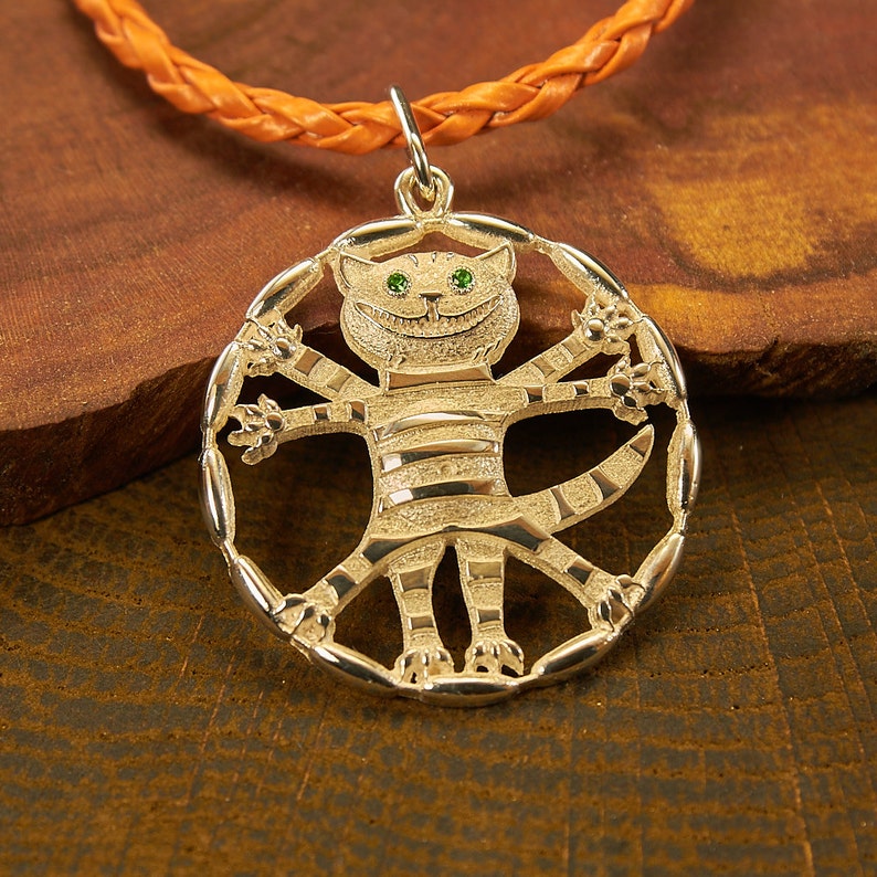 Peut inclure: Un collier avec un pendentif en argent repr&eacute;sentant le Chat du Cheshire d'Alice au Pays des Merveilles. Le chat est repr&eacute;sent&eacute; avec un large sourire et des yeux verts, ses bras et ses jambes &eacute;tant tendus dans un design circulaire.